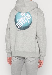 Sudadera con capucha gris con un estampado gráfico circular en verde azulado y rosa, con el texto "Levi's Silver Lab", sobre una tela suave y texturizada.