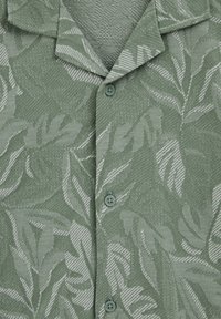 Chemise verte boutonnée avec un motif subtil de feuilles texturées et un col classique, présentée en gros plan.
