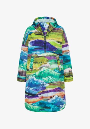 Femme debout contre un mur blanc, portant une veste à capuche colorée et oversize avec des motifs abstraits, un jean bleu et des baskets violet clair.