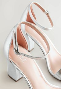 Sandales à talons carrés en argent avec une sangle de cheville réglable et une semelle intérieure lisse rose. Le matériau supérieur a une finition métallique avec un design épuré.
