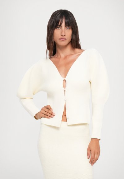 Bela pletena kardigan s globokim V-izrezom, napučenimi rokavi in sprednjim gumbom za zapiranje. Ima teksturirano tkanino in skrajšan dizajn.