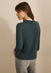 Tee-Farbenes Waffle-Strickoberteil mit rundem Ausschnitt und drei Viertel-Ärmeln, kombiniert mit hellgrauen Jeans. Weiche Textur und lockere Passform.