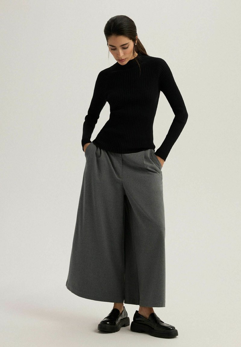 Czarny prążkowany sweter z golfem w połączeniu z szerokimi szarymi spodniami. Model stoi z rękami w kieszeniach, nosząc czarne buty. Minimalistyczny design.