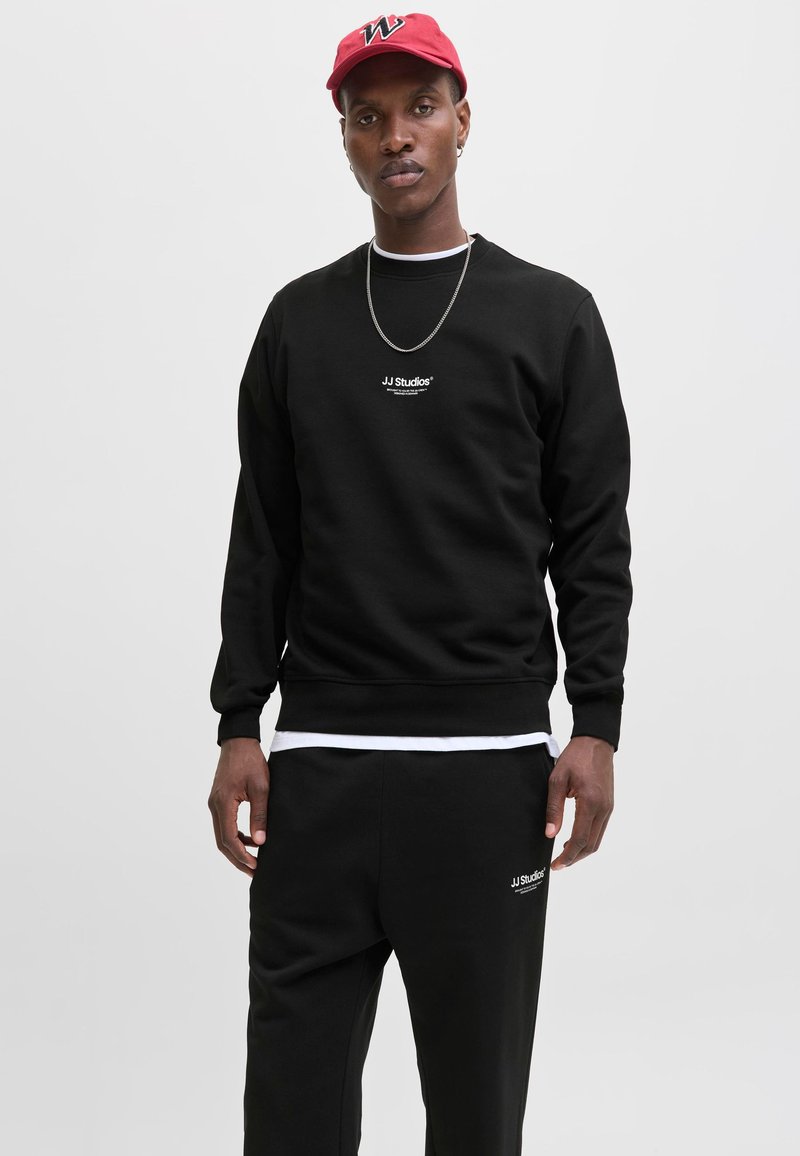 Sudadera negra con logo en el pecho, puños y dobladillo acanalados, combinada con pantalones negros a juego. Sombrero rojo con logo. Collar plateado como acento.