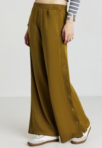 Pantalon ample en tissu vert olive avec des boutons pression sur les côtés, une taille élastique et une texture lisse, associé à des baskets blanches.