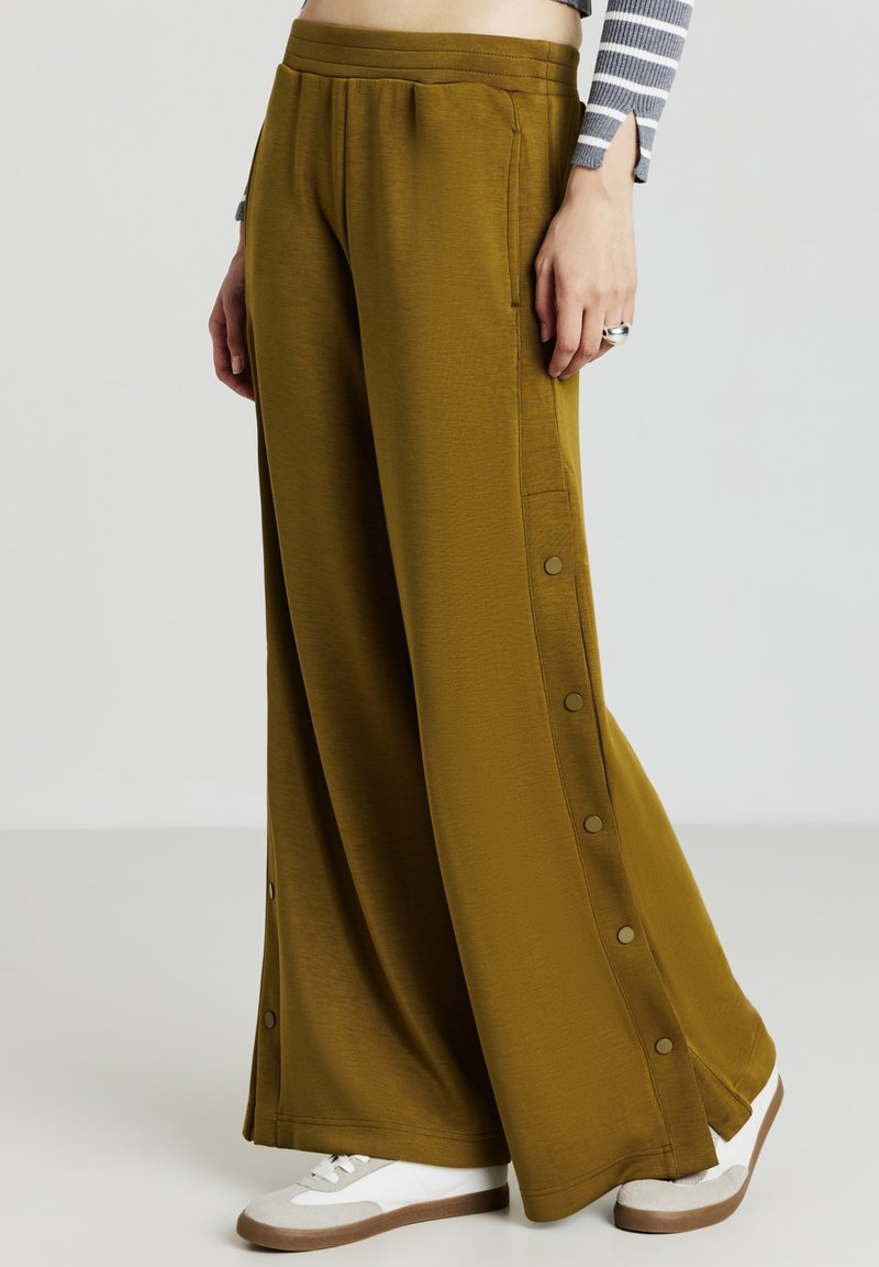 Pantalon ample en tissu vert olive avec des boutons pression sur les côtés, une taille élastique et une texture lisse, associé à des baskets blanches.