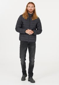 Polstret sort jakke med høj krave, der har kviltede sektioner. Parret med mørke jeans og grå ankelstøvler, stående på en hvid baggrund.