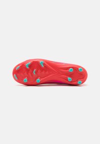Chaussure de football avec une semelle extérieure rouge, présentant une surface texturée, six crampons proéminents et des accents teal.