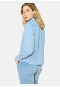 Camicia di jeans azzurro chiaro con colletto a punta, tasche sul petto e maniche lunghe. Presenta una vestibilità comoda e un orlo leggermente curvato.