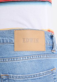 Blå denimjeans med en tanläderlapp märkt "EDWIN" på midjebandet. Vit skjorta med flerfärgade horisontella ränder delvis synlig.