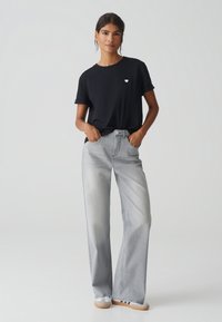 T-shirt di cotone nero con un piccolo logo a forma di cuore bianco, abbinato a jeans in denim a gamba larga grigio chiaro. Le scarpe presentano un design a righe bianche e blu.