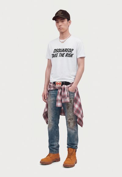 Jeune homme portant un t-shirt blanc "DSQUARED2 TAKE THE RISK", un jean usé, des bottes jaunes, une chemise à carreaux nouée autour de la taille, et une casquette noire.