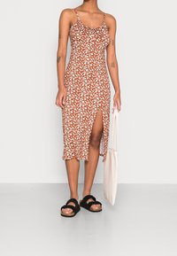Vestido midi marrón con estampado floral, tirantes finos, escote fruncido, abertura lateral, combinado con sandalias negras y un bolso de mano ligero.