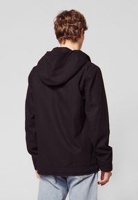 Czarna kurtka z kapturem o luźnym kroju, z gładką fakturą i widocznymi szwami, w połączeniu z jasnoniebieskimi dżinsami.