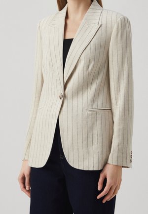 Femme portant un blazer beige à fines rayures avec un seul bouton sur un haut noir et un pantalon foncé, sur un fond uni.