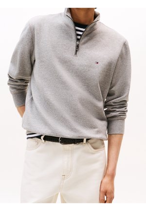 Mand iført lysegrå kvart-zip sweater over sort og hvid stribet skjorte, kombineret med hvide bukser og sort flettet bælte.