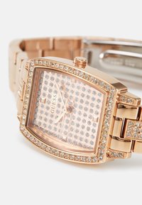 Rosaguld rektangulært ur med en krystalbesat bezel og urskive, sammenføjet rem og et minimalistisk ansigt, der viser brandnavnet "GUESS."