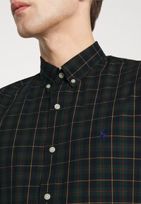 Skjorta med knappar i mörkgrön rutigt mönster med röda och gula linjer; har en klassisk krage, vita knappar och en liten blå logotyp på bröstet.