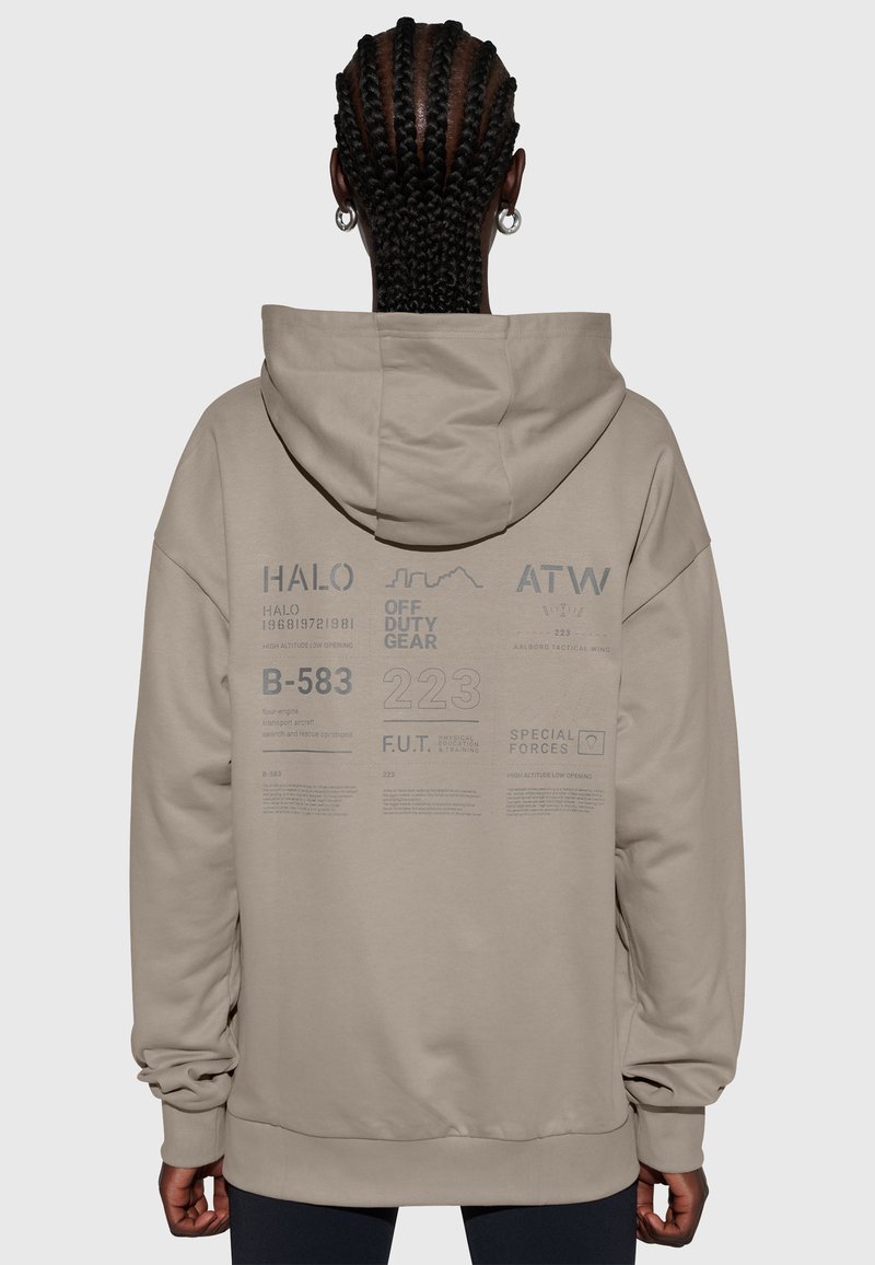 Osoba z warkoczami, nosząca beżową bluzę z kapturem z tekstem i logo, w tym "HALO", "OFF DUTY GEAR" i "SPECIAL FORCES" na plecach.