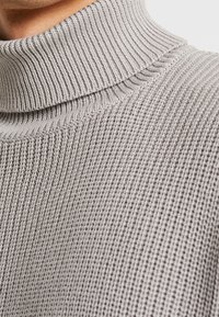 Nahaufnahme eines hellgrauen gestrickten Rollkragensweaters, der den gerippten Kragen und das strukturierte Muster des Stoffs zeigt.