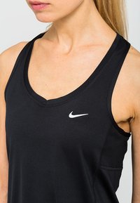 Černý sportovní top s hlubokým výstřihem ve tvaru lodi, na levém prsu se nachází malé bílé logo Nike a má přiléhavý střih. Hladká textura.