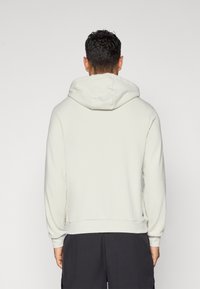 Mand med kort krøllet hår iført en lysegrå hættetrøje og sorte bukser, vender væk mod en ensfarvet baggrund.