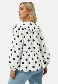Blusa bianca con grandi pois neri, colletto arricciato e maniche a sbuffo. Presenta un corpetto aderente con orlo peplum arricciato.