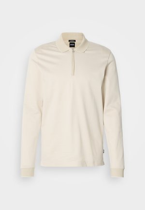 Polo a maniche lunghe di colore avorio, realizzato in tessuto textured. Presenta un colletto con zip a metà e dettagli in costina sui polsini.