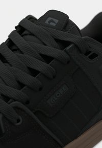 Κοντινό πλάνο μαύρου αθλητικού παπουτσιού Globe με χοντρές κορδόνια και καφέ σόλα, με ραμμένα λεπτομέρειες και λογότυπο της μάρκας στο πλάι και στη γλώσσα.