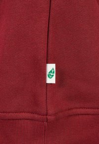 Roter Stoff mit einem kleinen weißen Etikett, das ein grünes Blattsymbol zeigt, das in eine Naht nahe der unteren Kante genäht ist.