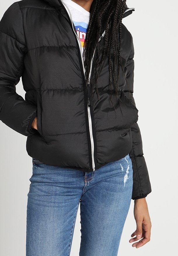 Femme portant une veste matelassée noire avec une fermeture éclair blanche, un jean bleu déchiré et un t-shirt blanc à motif, debout avec une main dans la poche.