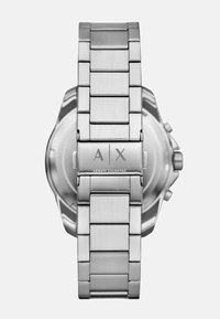 Srebrna ura iz nerjavečega jekla z brušenim zaključkom, zasnova iz povezovalnih členov in graviran logotip na zaponki; okrogla urina obraz, viden iz zadnje strani.