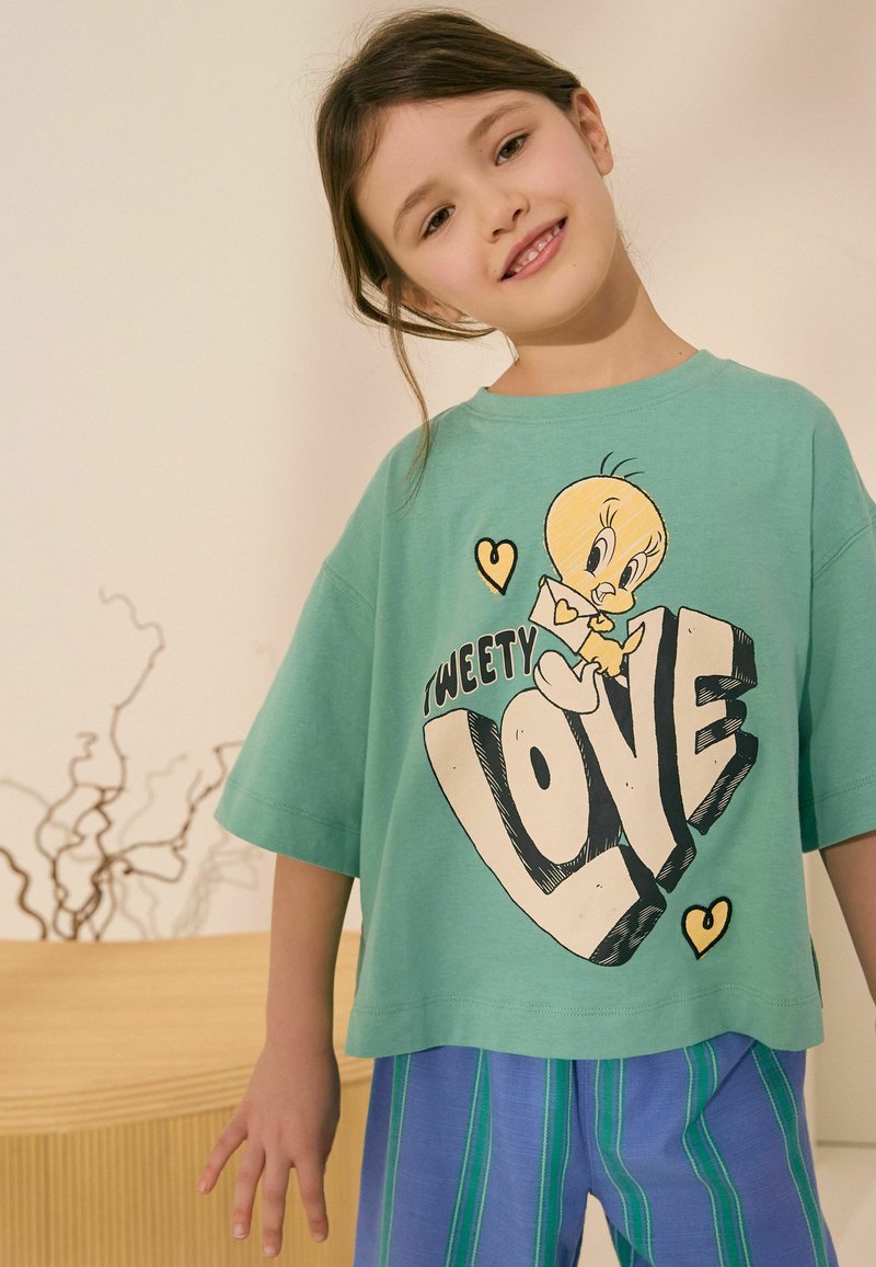 Ragazza giovane sorridente, indossa una T-shirt verde oversize con il personaggio di Tweety e il testo "LOVE", abbinata a pantaloni a righe blu e verdi.
