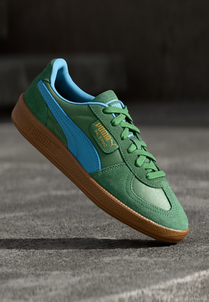 Baskets vertes Puma avec bande bleue et doublure intérieure, semelle marron et étiquette dorée "Puma Palermo", montrées flottant au-dessus d'une surface grise.