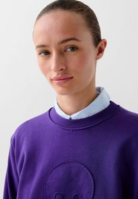 Paarse sweatshirt met een ronde halslijn, met een gestructureerd ontwerp. Draagt over een lichtblauwe blouse met opstaande kraag, wat de laagjes benadrukt.