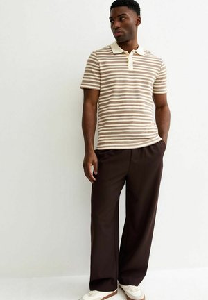 Homme debout portant un polo à manches courtes rayé beige et blanc, un pantalon large de couleur foncée et des baskets blanches sur un fond uni.