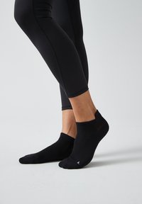 Schwarze, kurz geschnittene Socken aus einem strukturierten Material, mit einem gerippten Bündchen und einem kleinen weißen Etikett an der Seite. Getragen mit schwarzen Leggings.