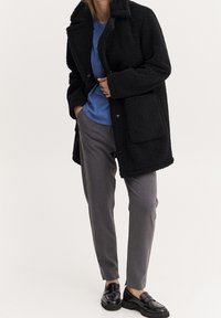 Zwarte, pluizige, gestructureerde jas met kraag en zakken aan de voorkant, gedragen over een blauw overhemd en grijze op maat gemaakte broek, gecombineerd met zwarte loafers.