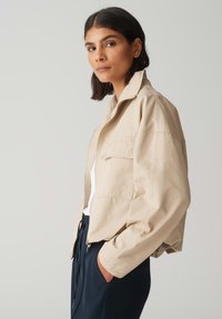 Beige cropped jack met een lichtgewicht stof, met een rits aan de voorkant, een puntige kraag en een borstzak. Geken op een witte top.