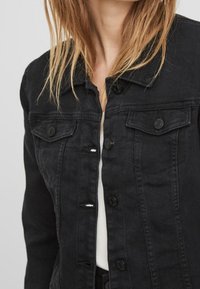 Svart denimjacka med klassisk krage, framknäppning med knappar och två bröstfickor. Har en texturerad tyg och en något figursydd design.