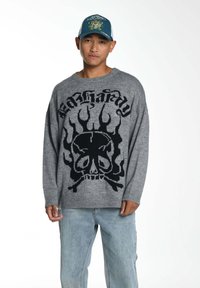 Grauer Strickpullover mit schwarzem Totenkopf- und Flammendesign, mit rundem Ausschnitt und gerippten Bündchen, kombiniert mit hellblauen Jeans.