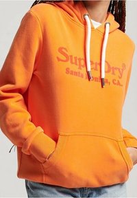Sudadera naranja con bolsillo canguro, capucha con cordón y puños acanalados. Presenta el logo "SuperDry" en rojo en el pecho.