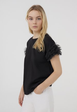 Maglietta nera a maniche corte con dettagli in tulle nero sovrapposti sulle spalle, indossata con pantaloni bianchi. Tessuto morbido, vestibilità rilassata.