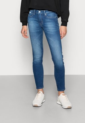 Jeans skinny en denim bleu moyen avec un léger délavage, design à cinq poches et fermeture classique par bouton et zip. Associés à des baskets blanches.