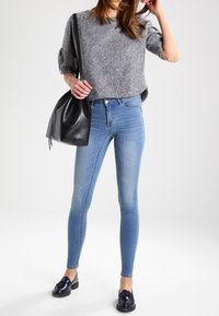 Siniset farkut, jotka on valmistettu denimistä, on yhdistetty harmaaseen lyhythihaiseen neulepaitaan ja mustaan laukkuun. Jalkineet ovat kiiltävät mustat loafer-kengät.