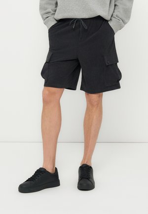 Άνδρας που φοράει μαύρα cargo σορτς με κουμπωτές πλαϊνές τσέπες, γκρι φούτερ και μαύρα αθλητικά παπούτσια, στέκεται μπροστά σε λευκό φόντο.