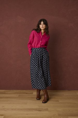 Femme portant un pull rose boutonné, un pantalon large bleu marine à pois et des bottines marron, debout contre un mur marron et un sol en bois.