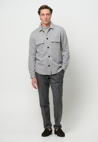 Grijze wollen shirtjack met knoopsluiting en twee borstzakken, gecombineerd met grijze wollen broek en donkerbruine suède instapschoenen.