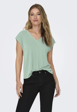 Camiseta de cuello en V de color verde menta con mangas cortas, hecha de material suave, combinada con jeans negros. La camiseta presenta un corte holgado y una textura sutil.