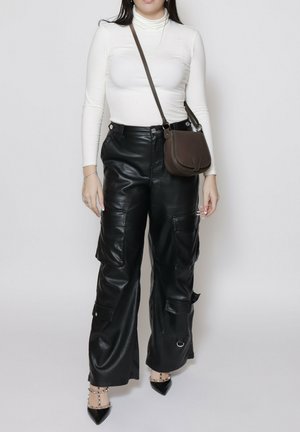 Schwarze Leder-Cargo-Hosen mit Seitentaschen, kombiniert mit einem eng anliegenden weißen Rollkragenshirt und einer braunen Umhängetasche. Spitz zulaufende schwarze Absätze betonen das Outfit.